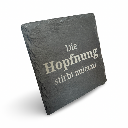 Schräge Ansicht eines Untersetzers aus Schiefer mit lasergraviertem Schriftzug „Die Hopfnung stirbt zuletzt"  – dekorativer Getränkeuntersetzer für Bierliebhaber